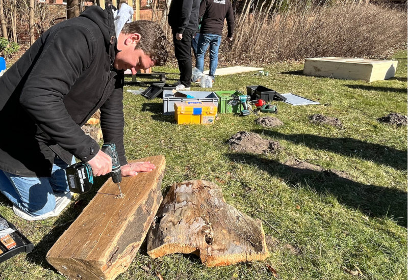 Nikolai Schade vom Coca-Cola-Team in Lüneburg bohrt unterschiedlich große Löcher in ein großes Stück Eichenholz. Er kniet auf dem Boden und hält einen Akku-Bohrer in der Hand. 