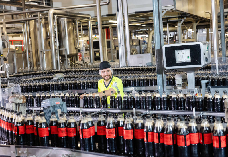 Mehrere PET-Flaschen mit Coca-Cola stehen auf dem Fließband in der Mehrwegabfüllung bei Coca-Cola in Fürstenfeldbruck. Ein Mitarbeitender steht an den Produktionsbändern und greift zu den Flaschen. 