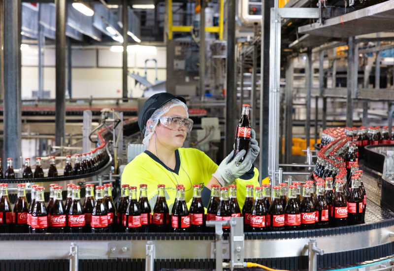 Eine Mitarbeitende an der Produktionslinie für Mehrweg-Glasflaschen bei Coca-Cola in Fürstenfeldbruck: Er hält eine Coca-Cola Glasflasche in der Hand, um sie zu prüfen.  