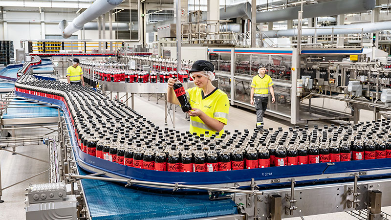 Eine Mitarbeiterin von Coca-Cola Karlsruhe steht in der Produktionshalle an einem Band, auf dem sich viele Flaschen Coca-Cola Zero Sugar befinden. Die Mitarbeiterin hat eine der Flaschen hochgenommen und betrachtet sie prüfend.