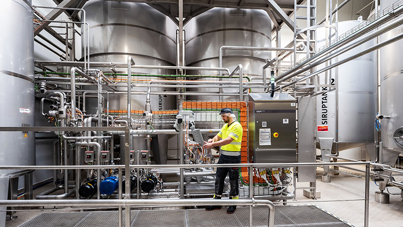 Der so genannte „Sirupraum“ des Coca-Cola Werks in Karlsruhe: Im Hintergrund hohe Tanks, im Vordergrund viele Rohre aus Edelstahl. Ein Mitarbeiter prüft einen Prozess auf einem kleinen Monitor. 