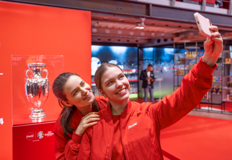 Mitarbeitende in guter Stimmung auf dem Coca-Cola Messestand machen Selfie mit Fußball-Pokal