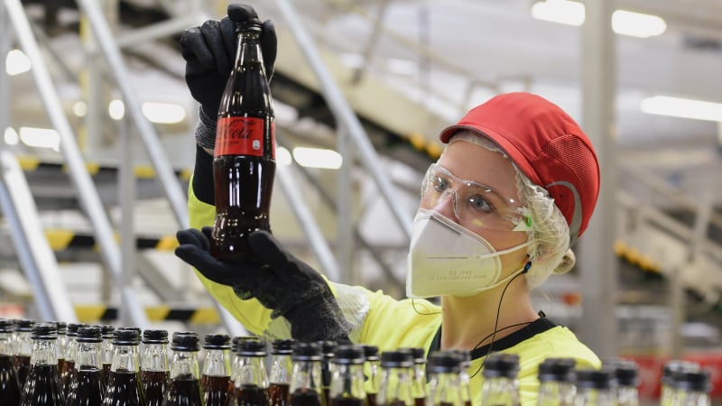 Eine Mitarbeiterin von Coca-Cola hält prüfend eine 0,4-Liter-Glasflasche mit Coca-Cola Zero Sugar hoch. Im Vordergrund ein Fließband mit mehreren Coca-Cola Flaschen. 