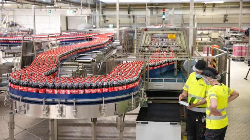 Die Produktionslinie mit abgefüllten und etikettierten Coca-Cola Flaschen ist in voller Größe zu sehen, daneben stehen zwei Mitarbeitende von Coca-Cola mit gelben Arbeitssicherheitsshirts, die gemeinsam etwas besprechen und auf Unterlagen schauen