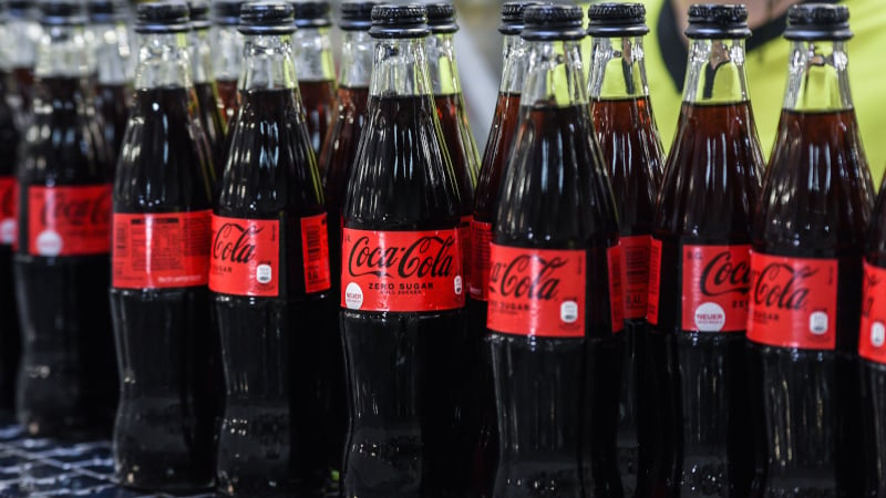 Mehrere Glasflaschen mit Coca-Cola Zero Sugar stehen auf dem Fließband in der Produktion. 