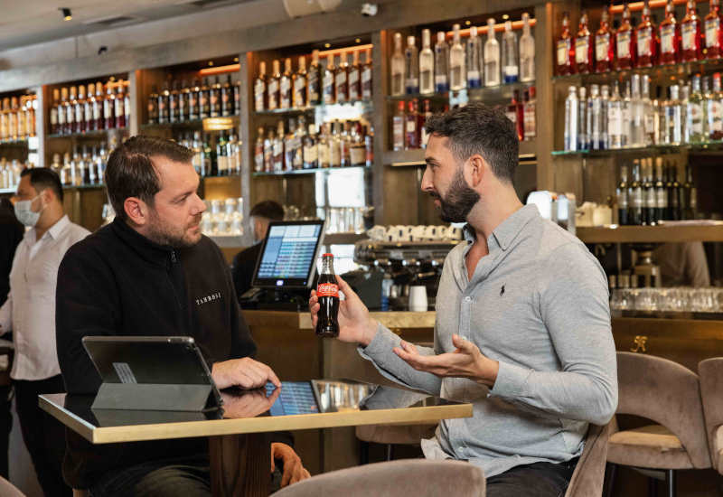 Ein Café-Mitarbeiter und ein Verkaufsberater von Coca-Cola sitzen im Café an einem Tisch. Der Verkaufsberater hält eine kleine Coca-Cola Flasche in der Hand. Die beiden Männer sprechen miteinander.