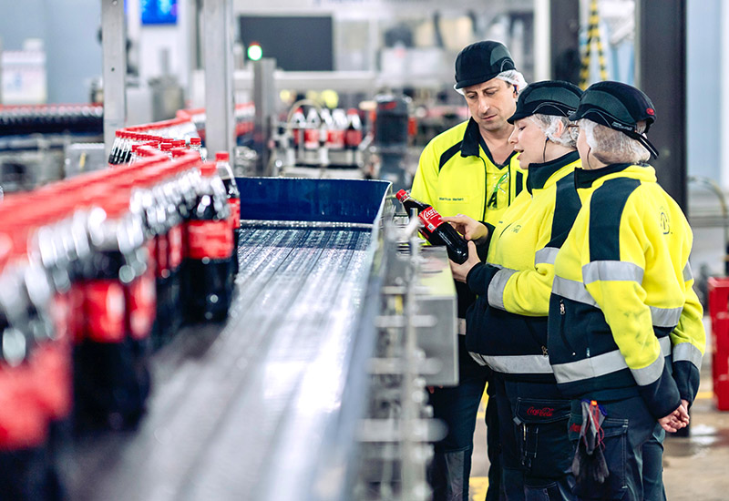 Coca-Cola Werk in Mönchengladbach mit 3 Mitarbeitenden vor dem Produktionsband