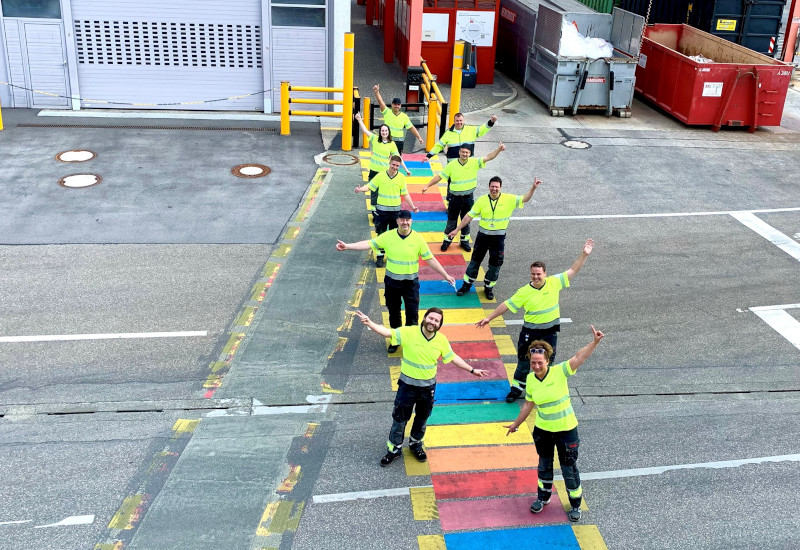 Regenbogen-Zebrastreifen auf dem Werksgelände bei Coca-Cola in Fürstenfeldbruck im Jahr 2021: Mitarbeitende vom Standort stehen auf dem Zebrastreifen und freuen sich