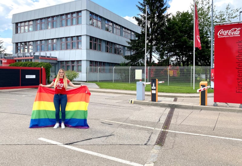 Foto von einer Mitarbeitenden am Standort in Fürstenfeldbruck. Sie hält eine Regenboggenflagge in der Hand und steht am Eingang des Werksgeländes.