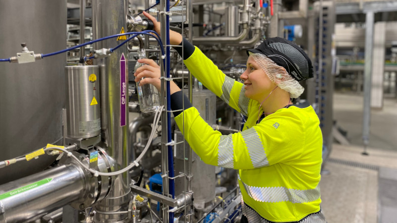 Coca-Cola Mitarbeiterin entnimmt Spülwasser am Produktionsstandort in Karlsruhe: Sie hält eine Flasche in der Hand, um sie herum sind Rohrleitungen. Sie trägt ein neongelbes Arbeitssicherheitsshirt, ein Haarnetz und eine Anstoßkappe.