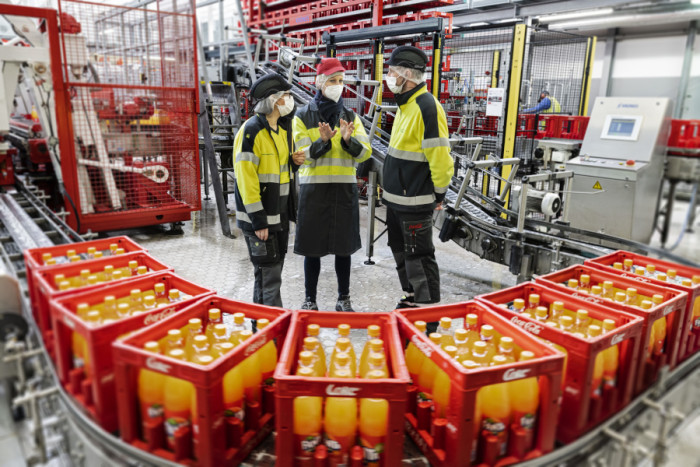 Coca-Cola Geschäftsführerin Personal, Kathrin Flohr, inmitten einer Produktionslinie von Coca-Cola im Gespräch mit zwei Kollegen, die neben ihr stehen. Im Vordergrund sind Mehrweg-Getränkekisten mit Fanta zu sehen. 