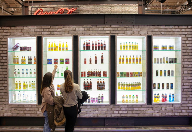 Zwei junge Frauen stehen vor einer Wand mit fünf beleuchteten Vitrinen, in denen Flaschen mit Getränken aus dem Hause Coca-Cola Europacific Partners zu sehen sind. 