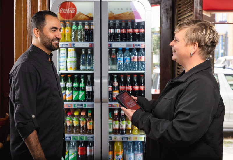 Ein Mann und eine Frau stehen vor einem Kühler mit Getränken aus dem Hause Coca-Cola Europacific Partners und sind in ein Verkaufsgespräch vertieft. 